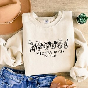 May include: Cream-colored sweatshirt featuring a black graphic of Mickey Mouse and friends, with the text "Mickey & Co. Est. 1928." The sweatshirt is paired with blue denim shorts, a tan hat, and sandals.