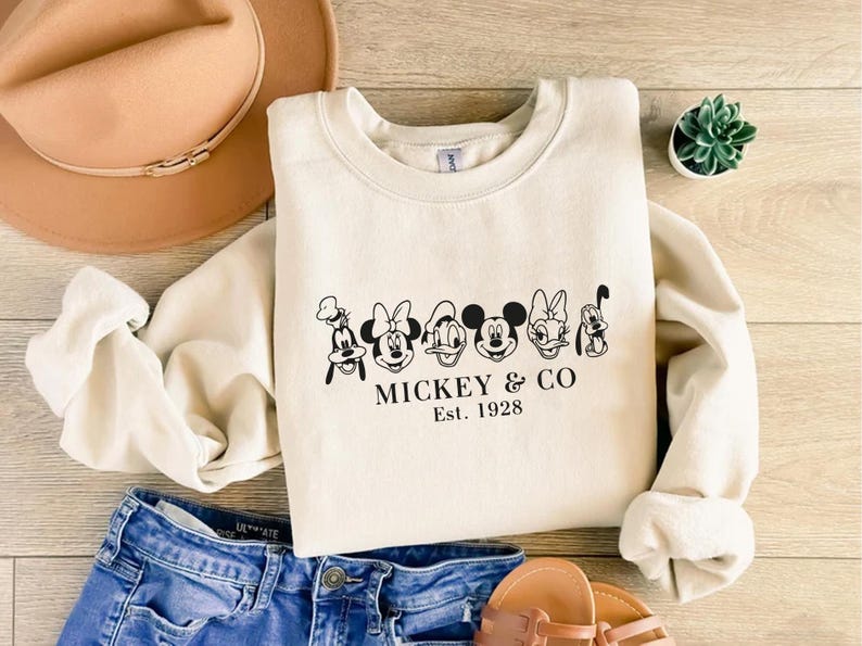 May include: Cream-colored sweatshirt featuring a black graphic of Mickey Mouse and friends, with the text "Mickey & Co. Est. 1928." The sweatshirt is paired with blue denim shorts, a tan hat, and sandals.
