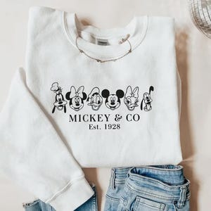 May include: White sweatshirt featuring a black and white graphic of Mickey Mouse, Minnie Mouse, Donald Duck, Daisy Duck, Goofy, and Pluto. The text "Mickey & Co. Est. 1928" is printed below the characters. A necklace is draped on the sweatshirt.
