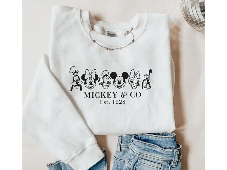 May include: White sweatshirt featuring a black and white graphic of Mickey Mouse, Minnie Mouse, Donald Duck, Daisy Duck, Goofy, and Pluto. The text "Mickey & Co. Est. 1928" is printed below the characters. A necklace is draped on the sweatshirt.