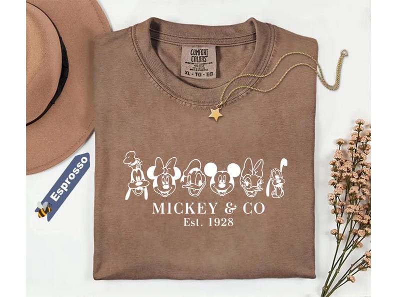 May include: Brown t-shirt featuring white cartoon character faces and the text "Mickey & Co. Est. 1928". A gold star pendant necklace and a brown hat are also visible. The shirt is a Comfort Colors brand.