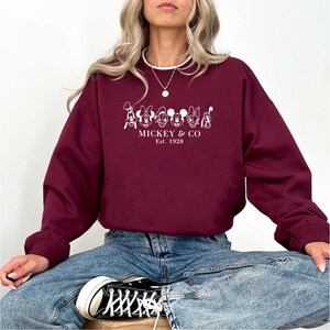 May include: A maroon sweatshirt featuring a white graphic of cartoon characters and the text "Mickey & Co. Est. 1928." The sweatshirt is long-sleeved and has a crew neck. The person is wearing blue jeans and black and white sneakers.