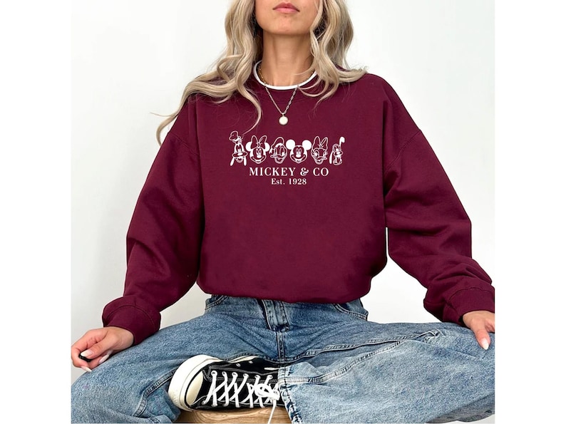 May include: A maroon sweatshirt featuring a white graphic of cartoon characters and the text "Mickey & Co. Est. 1928." The sweatshirt is long-sleeved and has a crew neck. The person is wearing blue jeans and black and white sneakers.