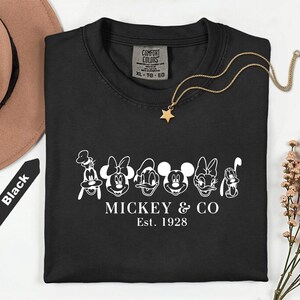 May include: Black t-shirt featuring a white graphic of Mickey & Co. characters with the text "MICKEY & CO Est. 1928". A gold necklace with a star pendant and a brown hat are also visible.