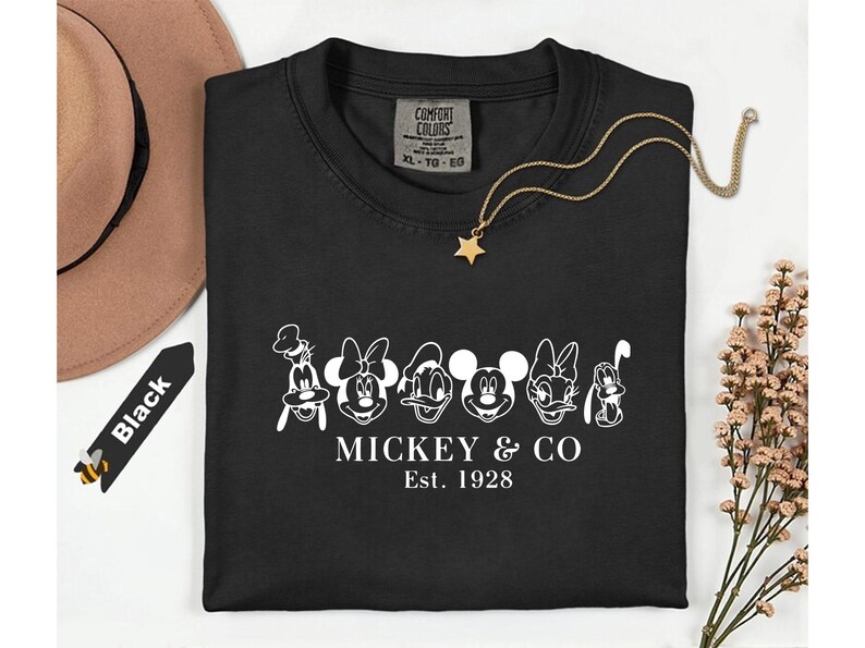 May include: Black t-shirt featuring a white graphic of Mickey & Co. characters with the text "MICKEY & CO Est. 1928". A gold necklace with a star pendant and a brown hat are also visible.