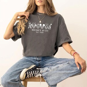 May include: Dark gray t-shirt with a white graphic of cartoon characters and the text "MICKEY & CO Est. 1928". The shirt is worn with blue jeans and black sneakers.
