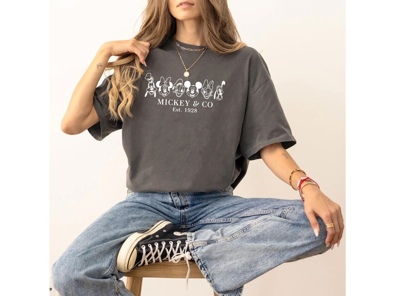 May include: Dark gray t-shirt with a white graphic of cartoon characters and the text "MICKEY & CO Est. 1928". The shirt is worn with blue jeans and black sneakers.