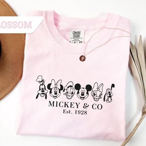 May include: A light pink t-shirt featuring a black graphic of Disney characters, including Mickey Mouse, Minnie Mouse, and Goofy. The text "MICKEY & CO Est. 1928" is printed below the characters. A gold necklace with a pendant is on the shirt.