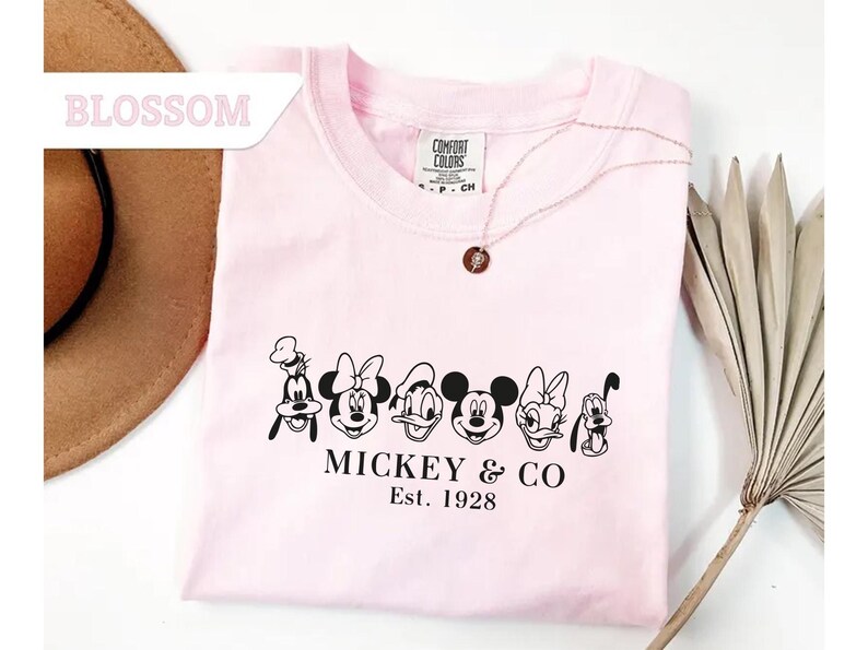 May include: A light pink t-shirt featuring a black graphic of Disney characters, including Mickey Mouse, Minnie Mouse, and Goofy. The text "MICKEY & CO Est. 1928" is printed below the characters. A gold necklace with a pendant is on the shirt.