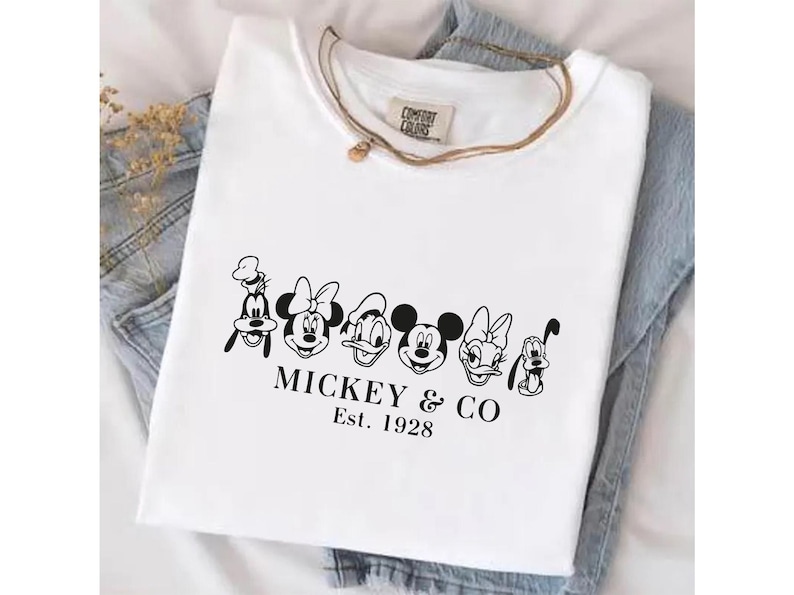May include: White t-shirt featuring a black graphic of Mickey Mouse and friends, including Goofy, Minnie Mouse, Donald Duck, Daisy Duck, and Pluto. The text "MICKEY & CO Est. 1928" is printed below the characters. The shirt is folded on denim.