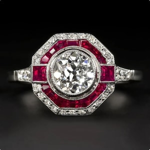 Pode incluir: Um anel de estilo vintage com uma grande pedra central de diamante transparente e redondo. O anel apresenta um design geométrico com uma borda de rubis vermelhos e diamantes menores. O anel é cravejado em um metal prateado.