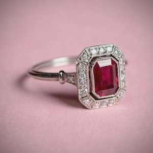 May include: A silver ring with a rectangular, deep red gemstone at the center, surrounded by smaller, clear stones. The ring has an ornate, vintage-inspired design, set against a soft pink background.