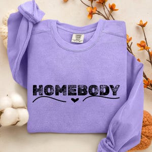 May include: A light purple sweatshirt with the word "HOMEBODY" in black block letters. A black heart is below the word. The sweatshirt is a crew neck style with long sleeves.