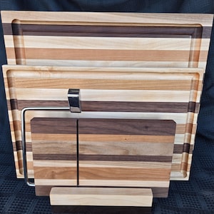 May include: A set of three wooden cutting boards and a cheese slicer. The boards are rectangular with a groove around the edges and are made of alternating strips of light and dark wood. The cheese slicer has a stainless steel wire and handle.