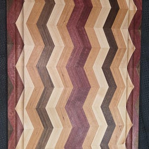 May include: A rectangular wooden chopping board with a chevron pattern. The board has alternating stripes of light, medium, and dark brown wood, with a central purple stripe. A carved groove runs around the edge.