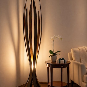 May include: A striking wooden floor lamp with a distinctive, open design. The lamp has vertical wooden slats that curve inwards at the top, forming a sculptural shape. Warm light shines from within, lighting the area. A small side table and a white sofa are also visible.