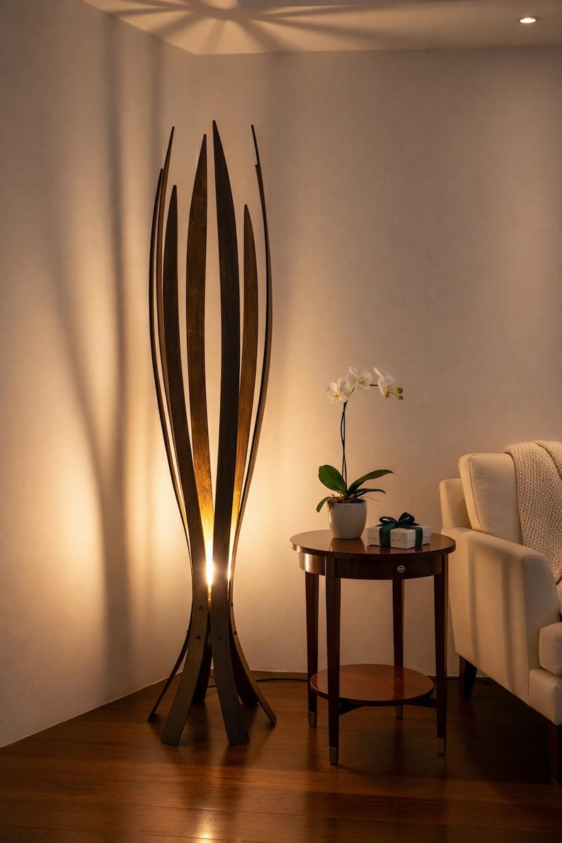 May include: A striking wooden floor lamp with a distinctive, open design. The lamp has vertical wooden slats that curve inwards at the top, forming a sculptural shape. Warm light shines from within, lighting the area. A small side table and a white sofa are also visible.