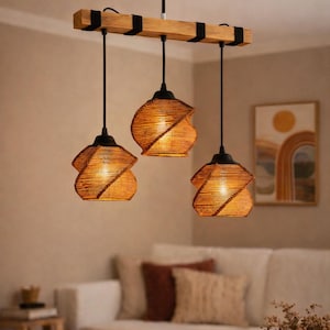 Könnte beinhalten: Eine Pendelleuchte mit drei Lichtern. Die Leuchte verfügt über drei gewebte, geometrisch geformte Schirme, die an einem Holzbalken aufgehängt sind. Die Schirme haben eine warme, natürliche Farbe, und der Balken ist hellbraun. Die Leuchte ist an einer schwarzen Deckenplatte befestigt.