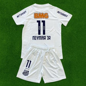 Peut inclure: Ensemble de maillot et short de football blancs. Le maillot porte le numéro 11, le nom "NEYMAR JR" et les logos "BMG" et "NETSHOES". Le short affiche également le numéro 11 et un logo d'équipe. L'ensemble est présenté sur un fond vert.