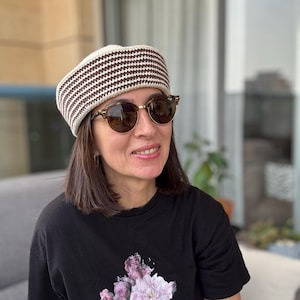 Op de afbeelding: Een crèmekleurige gehaakte hoed met een bruin-wit gestreept patroon. De hoed wordt gedragen met een ronde zonnebril met een gouden montuur en een zwart t-shirt met een bloemenmotief.
