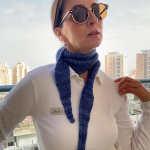 Può includere: Una sciarpa blu lavorata a maglia con un'estremità a punta è indossata intorno al collo. La sciarpa è di un blu intenso con accenti blu più chiari. La persona indossa occhiali da sole e una camicia bianca con colletto. Lo sfondo mostra edifici e un cielo luminoso.