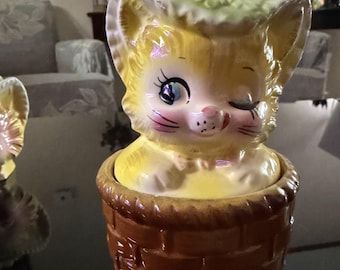 May include: A vintage ceramic cat figurine in a basket. The cat is yellow with blue eyes, pink cheeks, and a winking expression. It wears a hat with green floral accents. The basket is brown with a woven texture.