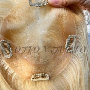 Puede incluir: Un postizo rubio con base de encaje y clips metálicos para sujetar. El texto "OTION STUDIO" es visible en la base. El cabello es de un color dorado claro.