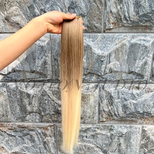 Puede incluir: Un mechón de extensiones de cabello rubio y castaño claro, sostenido contra un fondo de piedra gris. El cabello pasa de un marrón más oscuro en la parte superior a un rubio más claro en la parte inferior, creando un efecto ombre. El cabello parece liso y lacio.