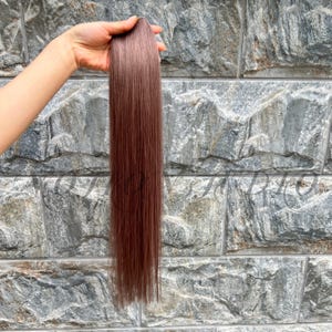 Könnte beinhalten: Ein Bündel glatter, rotbrauner Haarverlängerungen, das vor einer strukturierten Steinmauer gehalten wird. Das Haar hat eine einheitliche Farbe und wirkt glatt und glänzend. Die Extensions sind lang und scheinen aus Kunst- oder Echthaar zu bestehen.