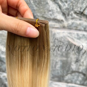 以下が含まれることがあります： 茶色からブロンドへのグラデーションのヘアエクステンション。髪は金色の留め金で織り込まれています。「NOTION STUDIO」の文字が背景に見えます。