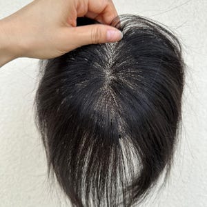 May include: A dark brown hair topper with a fine, lace-like base. The hair is held by a hand, showcasing the natural texture and color. The hairpiece appears to be designed for adding volume or coverage.