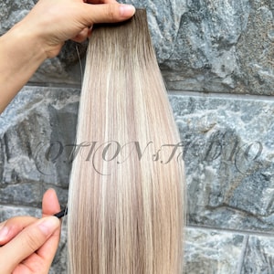 Puede incluir: Un mechón de extensiones de cabello con una raíz marrón oscuro que se desvanece en mechones rubios y marrón claro. El cabello es sostenido por una mano contra un fondo de piedra texturizada. El texto "MOTION STUDIO" es visible en el fondo.