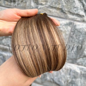 以下が含まれることがあります： 茶色とブロンドのハイライトが入ったヘアエクステンションの束。髪は手で持たれており、画像の下部に「MOTTO DIO」というテキストが表示されています。髪は滑らかでストレートに見えます。