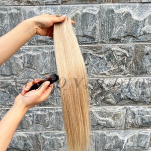 Puede incluir: Un mechón de extensiones de cabello rubio con reflejos, sostenido contra un fondo de piedra gris texturizada. Un peine negro se sostiene cerca del cabello. El cabello parece liso y lacio, con diferentes tonos de rubio.