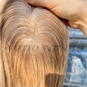 Puede incluir: Primer plano de una peluca rubia clara con una línea de cabello de aspecto natural. El cabello es liso y tiene una sutil mezcla de colores, con raíces más oscuras y puntas más claras. La peluca parece estar hecha de fibras sintéticas de alta calidad.