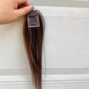 Könnte beinhalten: Eine braune Haarverlängerung mit Clip-Befestigung. Das Haar ist dunkelbraun und scheint glatt zu sein. Der Clip ist aus Metall und an einem kleinen, quadratischen Stoffstück befestigt.