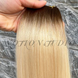 Puede incluir: Primer plano de una extensión de cabello con un efecto ombre marrón oscuro a rubio. El cabello es liso y parece ser cabello humano. El texto "MOTION STUDIO" es visible en la imagen.