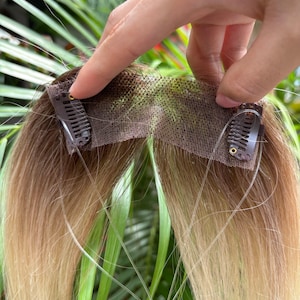 Puede incluir: Una extensión de cabello con un degradado de color de marrón a rubio. La sección superior consiste en una base de encaje fino de color beige con dos pequeñas pinzas metálicas de color marrón oscuro sujetas a los lados para fijación.