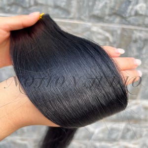 Puede incluir: Un mechón de extensiones de cabello negro liso sostenido en una mano. El cabello parece suave y liso, con un ligero brillo. Los extremos están cuidadosamente agrupados con una banda dorada. El fondo es una superficie gris texturizada.