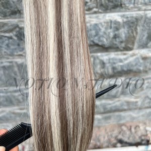 以下が含まれることがあります： 明るいブロンド、茶色、白のストライプが交互に並んだヘアエクステンションのクローズアップ。黒い櫛が髪に差し込まれています。「NOTION STUDIO」の文字が背景に見えます。