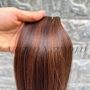 Puede incluir: Un mechón de extensiones de cabello castaño con reflejos castaños claros y cobrizos. El cabello es liso y lacio, con un brillo natural. Las extensiones se mantienen juntas en la parte superior con un hilo amarillo.