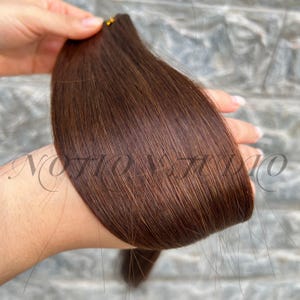 Könnte beinhalten: Ein Bündel dunkelbrauner Haarverlängerungen. Das Haar ist glatt und gerade, mit leichtem Glanz. Das Haar wird in einer Hand gehalten, wobei die Haarspitzen sichtbar sind.