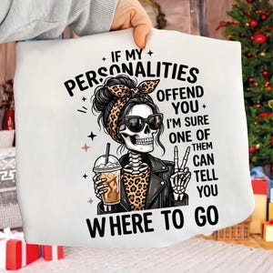 May include: White graphic tee featuring a skeleton with sunglasses, a leopard print top, and a leather jacket, holding a coffee. The text reads: "If my personalities offend you, I'm sure one of them can tell you where to go."