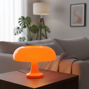 Puede incluir: Una lámpara de mesa naranja con forma de hongo, con una pantalla redondeada y una base de pedestal. La lámpara es de material brillante y está sobre una mesa de madera marrón oscuro. Un sofá y una planta de interior están en el fondo.