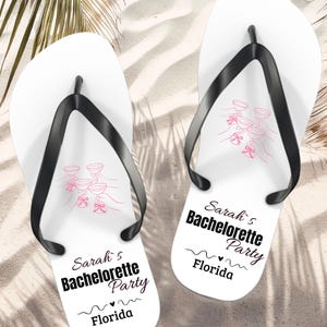 May include: White flip-flops with black straps, featuring pink illustrations of champagne glasses and text that reads "Sarah's Bachelorette Party Florida". The flip-flops are on a sandy surface.