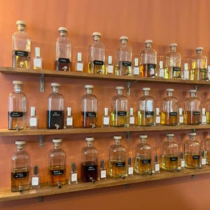 Puede incluir: Una colección de frascos de perfume de vidrio transparente con tapones de corcho y grifos de metal dispuestos en tres estantes de madera. Cada botella grande presenta una etiqueta rectangular negra con texto blanco, intercalada con frascos más pequeños.