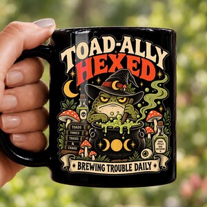 May include: A black ceramic mug featuring a colorful graphic of a toad wearing a witch hat, stirring a bubbling green cauldron. The text reads TOAD-ALLY HEXED, TOADS TONICS TRICKS & CHAOS, GOOD SPELLS BAD DECISIONS, and BREWING TROUBLE DAILY.