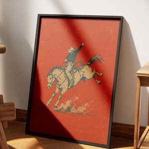 May include: A framed rectangular wall art print featuring a dark red background with a distressed illustration of a cowboy in a hat riding a bucking horse. The rider has one arm raised, and a coiled lasso hangs from the saddle of the tan horse.