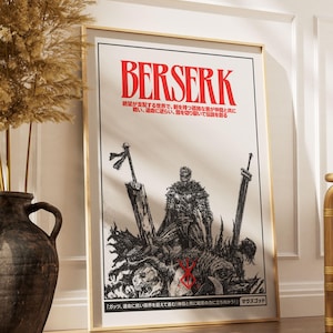 May include: A framed poster with the word "BERSERK" in red, above Japanese text. The black and white illustration depicts a warrior with swords and a graphic scene. The poster is in a gold frame, leaning against a wall.