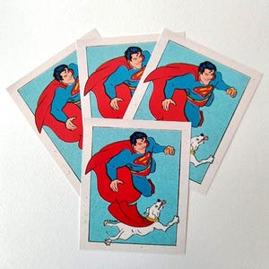 May include: Four vintage Superman trading cards. Each card shows Superman in a blue suit and red cape, flying with a white dog. The background is light blue, and the cards have white borders.